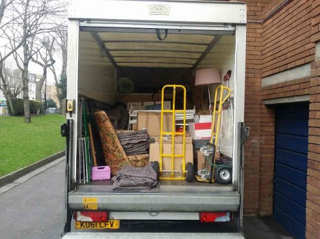 Photograph of the open rear of a white commercial moving truck parked on a driveway beside a brick building, with a green lawn and a few leafless trees visible in the background. The truck's interior is filled with various pieces of household and office rubbish, including a patterned upholstered armchair leaning against the left side, partially covered with a fabric sheet. In front of the armchair, there is a black and brown folded blanket or mat, and a pink plastic container towards the bottom left. A yellow hand trolley is positioned centrally inside the truck, with its base resting on the floor. To the right of the trolley, there are several cardboard boxes stacked together, some with white and red markings, and a brown lampshade sits atop a piece of furniture or cardboard. On the far right, a black and silver portable heater or similar device is partially visible. The truck’s interior ceiling has a mounted hook, and the overall scene depicts a typical collection of bulk household waste prepared for disposal or removal by Commercial Waste Erith, reflecting a cluttered arrangement suitable for rubbish removal services in the local area near Erith.