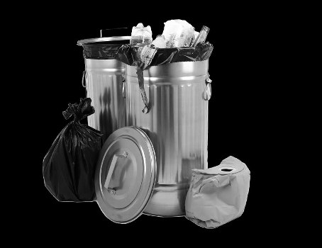 A large, cylindrical stainless steel rubbish bin with a closed lid, positioned on a black background. The bin contains visibly overflowing waste, including crumpled paper, plastic bags, and other household rubbish that extends above the rim. To the left of the bin is a tied black plastic rubbish bag leaning against its side, and on the right side is a smaller paper bag or parcel. The scene suggests a general waste collection setup, typical of a waste management service in the Erith area, with attention to waste disposal and rubbish removal. The polished surface of the bin contrasts with the crumpled textures of the bags and waste materials, highlighting the contrast between clean containers and disorderly rubbish, which Commercial Waste Erith aims to manage efficiently.