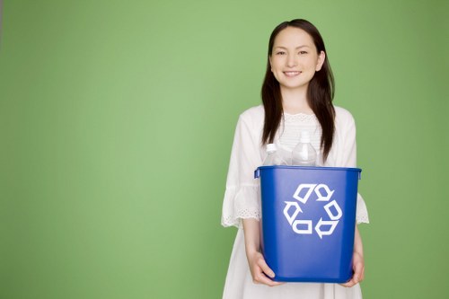 A young woman with long dark hair and a friendly expression is standing against a plain green background, holding a blue recycling bin with a white recycling symbol on the front. She is wearing a white, short-sleeved dress with lace details on the sleeves. The bin is positioned at waist height, and she is gripping it with both hands, one on each side. The environment is minimalistic, emphasizing the bin and her presentation of it, in a setting that suggests awareness of waste segregation and recycling practices. This scene subtly relates to rubbish and waste management services, such as those provided by Commercial Waste Erith, and highlights the importance of recycling in the context of proper waste disposal and environmental responsibility, particularly relevant to the Erith area in London postcode SE or DA. The lighting is soft and even, giving the image a clean and professional appearance suitable for online content about waste management services.