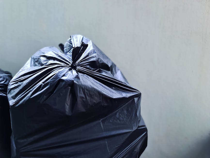 A collection of overflowing rubbish bins and scattered waste bags on a paved urban street corner with a low metal railing nearby. The central large grey bin is filled with mixed paper and cardboard, w