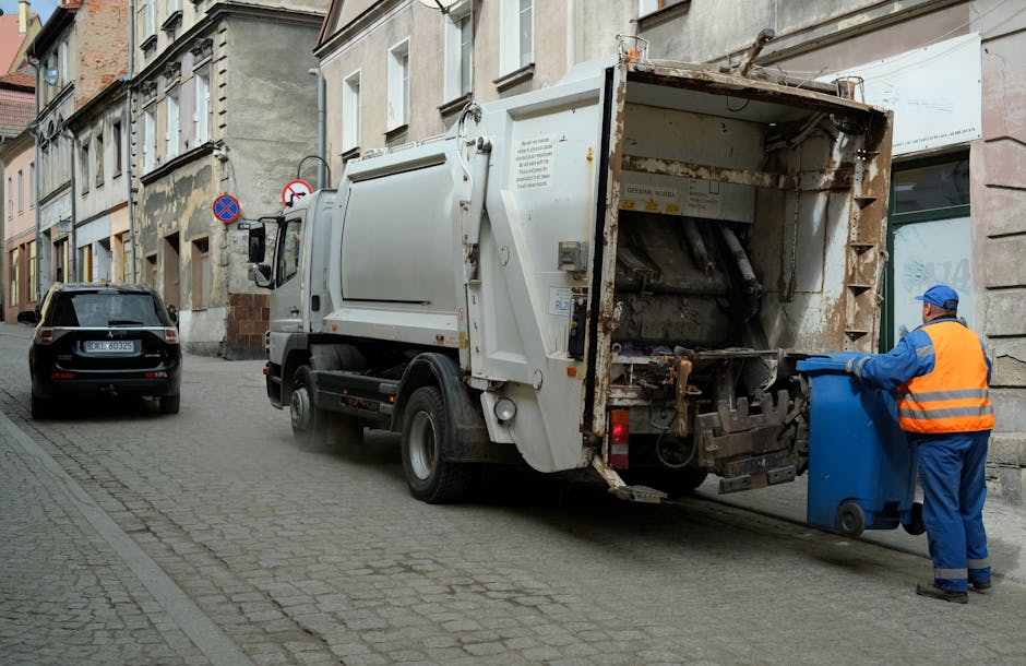 A worker wearing a high-visibility yellow vest and dark clothing operates a rubbish collection vehicle on an urban street during dusk or early evening. The worker, seen from behind, is using a control