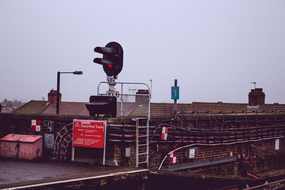 A bus shelter situated at a train station platform, featuring six white plastic seats with metal frames arranged in two groups of three. The shelter has a black metal roof supported by vertical black